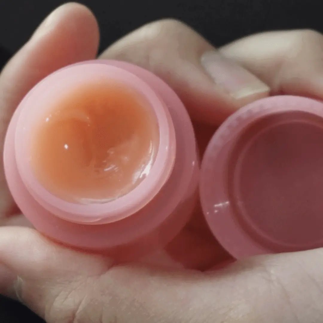Pink lip balm container with a visible product inside, held in a hand against a dark background.